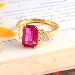 Op de afbeelding: Een gouden ring met een grote, roze, smaragd geslepen edelsteen omringd door kleinere, heldere edelstenen.