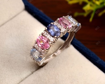 Puede incluir: Un anillo de banda de plata con una fila de piedras preciosas ovaladas. Las piedras preciosas son rosas, azules y azul claro. El anillo está engastado con pequeños diamantes.