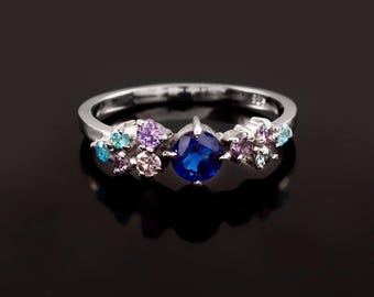 Puede incluir: Un anillo de plata con una piedra preciosa azul rodeada de piedras preciosas más pequeñas de color rosa, morado y azul.