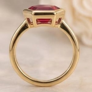 May include: A gold ring featuring a vibrant, deep red, emerald-cut gemstone set in a polished bezel setting. The band is smooth and circular, crafted from a warm-toned yellow metal that complements the faceted red stone held securely at the top.