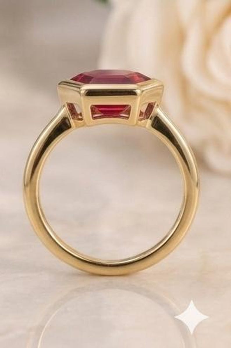 May include: A gold ring featuring a vibrant, deep red, emerald-cut gemstone set in a polished bezel setting. The band is smooth and circular, crafted from a warm-toned yellow metal that complements the faceted red stone held securely at the top.