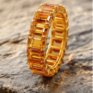 May include: An eternity band ring featuring a continuous row of emerald-cut yellow-orange gemstones set in a polished gold metal frame. The ring displays a repeating pattern of rectangular stones held by small metal prongs, creating a uniform design.