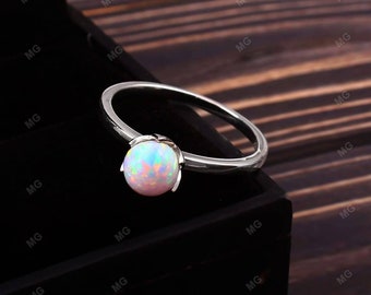 Pode incluir: Um anel de tom prateado com um cabochão de opala redondo e iridescente, fixado numa montagem de quatro garras. A pedra preciosa exibe reflexos de luz rosa, azul e verde sobre uma base branca leitosa, numa banda de metal lisa e polida.