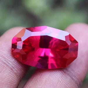 Op de afbeelding: Een gefacetteerde, levendige rode edelsteen met een achthoekige vorm. De edelsteen wordt in een hand gehouden, waarbij de facetten het licht reflecteren. De kleur van de edelsteen is een diep, rijk rood, met een gepolijst oppervlak.