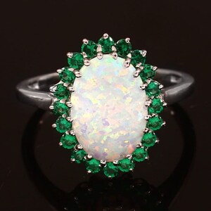 Puede incluir: Un anillo de plata con una gran opala blanca rodeada por un halo de piedras preciosas verdes.