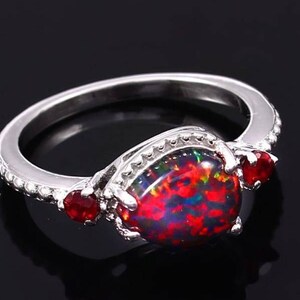 May include: A silver ring with a large, teardrop-shaped black opal gemstone surrounded by two smaller red gemstones.