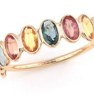 May include: A gold band ring featuring seven oval-shaped gemstones. The gemstones are arranged in a rainbow pattern, with blue, green, yellow, orange, and red stones.