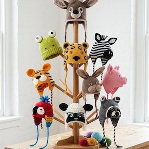 May include: A collection of handmade crochet animal hats, featuring a deer, frog, leopard, zebra, tiger, rabbit, pig, parrot, sheep, and raccoon. The hats are displayed on a wooden rack and table, showcasing the variety of colours and designs.