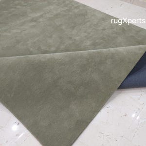 May include: A sage green rug with a soft, textured surface. The corner of the rug is folded back, revealing a dark blue backing. The rug is displayed on a light-colored tiled floor. The text "rugXperts" is visible.
