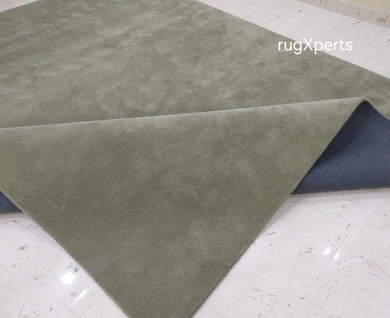 May include: A sage green rug with a soft, textured surface. The corner of the rug is folded back, revealing a dark blue backing. The rug is displayed on a light-colored tiled floor. The text "rugXperts" is visible.