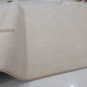 May include: A large, beige rug with a rolled-up edge. The rug has a soft, plush texture and appears to be made of a thick material. The brand name "rugXperts" is visible on the rug's surface. The rug is on a white floor.