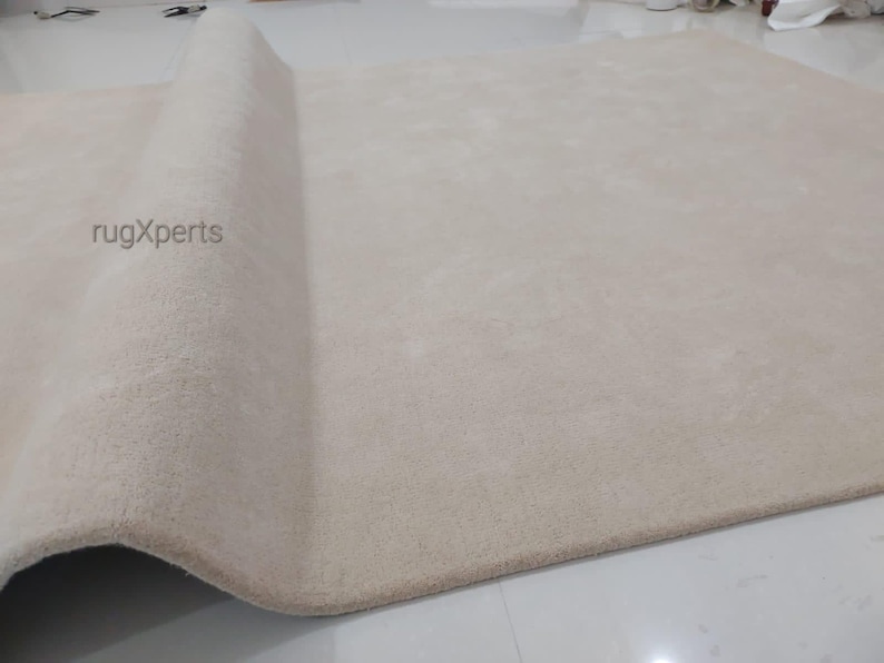 May include: A large, beige rug with a rolled-up edge. The rug has a soft, plush texture and appears to be made of a thick material. The brand name "rugXperts" is visible on the rug's surface. The rug is on a white floor.