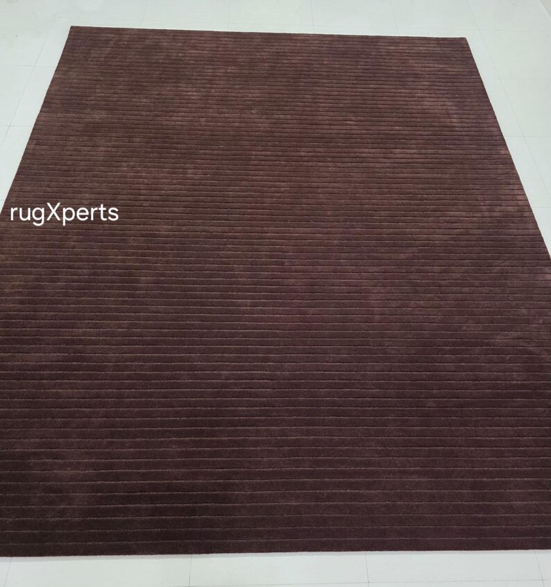 Op de afbeelding: Een rechthoekig bruin tapijt met een geribbelde textuur. Het tapijt heeft horizontale lijnen over het oppervlak. De tekst "rugXperts" is zichtbaar in de linker benedenhoek. Het tapijt ligt op een witte ondergrond.