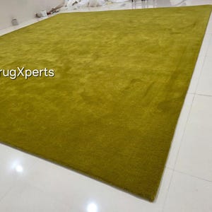 May include: A large, rectangular, olive-green rug. The rug has a soft, textured appearance and is placed on a white tiled floor. The text "rugXperts" is visible in the image.