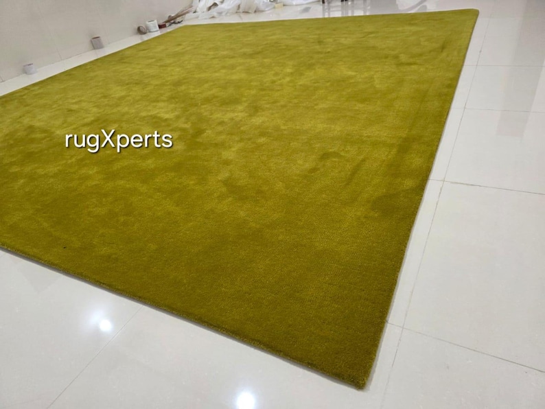 May include: A large, rectangular, olive-green rug. The rug has a soft, textured appearance and is placed on a white tiled floor. The text "rugXperts" is visible in the image.