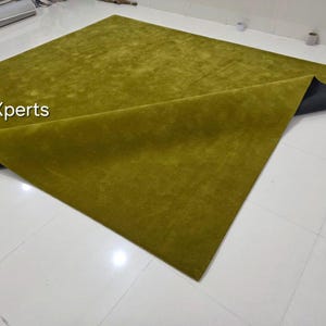 May include: A large, rectangular olive green rug with a soft, textured surface. The rug is partially folded, revealing a dark grey backing. The text "rugXperts" is visible in the upper left corner. The rug is placed on a white tiled floor.