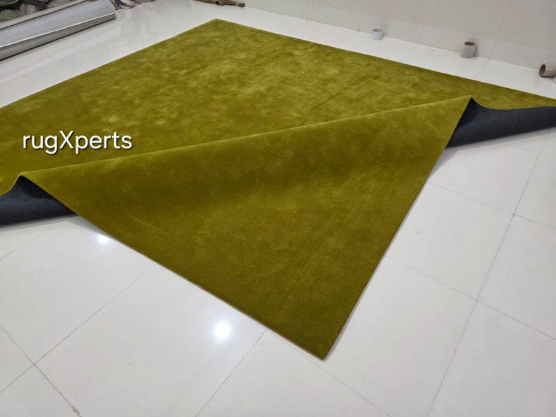 May include: A large, rectangular olive green rug with a soft, textured surface. The rug is partially folded, revealing a dark grey backing. The text "rugXperts" is visible in the upper left corner. The rug is placed on a white tiled floor.