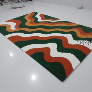 May include: A rectangular rug with a wavy pattern in green, orange, white, and rust colors. The rug has a modern design and is suitable for living rooms or bedrooms. The text "rugXperts" is visible on the rug.