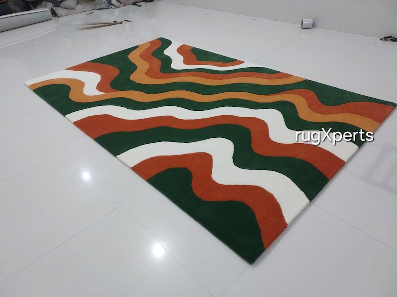 May include: A rectangular rug with a wavy pattern in green, orange, white, and rust colors. The rug has a modern design and is suitable for living rooms or bedrooms. The text "rugXperts" is visible on the rug.