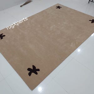 May include: A rectangular beige rug with four dark brown floral designs at each corner. The rug is placed on a white tiled floor. The text "rugXperts" is visible in the upper left corner.