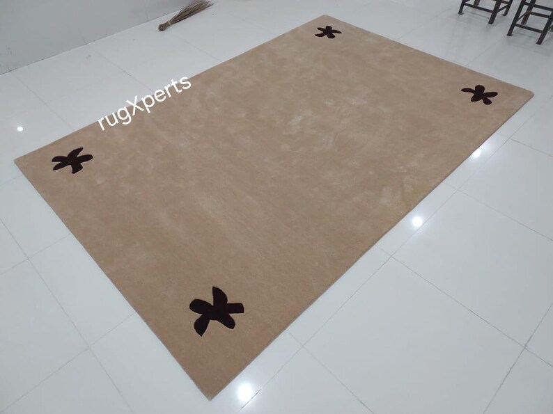 May include: A rectangular beige rug with four dark brown floral designs at each corner. The rug is placed on a white tiled floor. The text "rugXperts" is visible in the upper left corner.