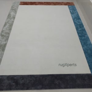 May include: A rectangular rug featuring a cream central area framed by a multi-coloured border. The border incorporates sections of grey, brown, blue, and dark grey. The rug's design is contemporary, suitable for home interiors.