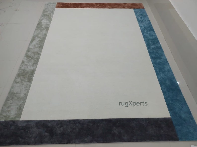 May include: A rectangular rug featuring a cream central area framed by a multi-coloured border. The border incorporates sections of grey, brown, blue, and dark grey. The rug's design is contemporary, suitable for home interiors.
