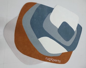 Puede incluir: Alfombra de área abstracta con un diseño de múltiples capas en tonos de marrón, blanco y azul. La alfombra tiene una forma única y orgánica y presenta una textura sutil. El texto "rugXperts" es visible en la esquina inferior derecha.
