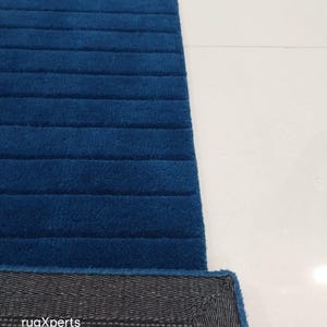 Op de afbeelding: Een gedeelte van een blauw tapijt met een geribbelde textuur. De rand van het tapijt onthult een grijze achterkant met de tekst "rugXperts". Het tapijt ligt op een wit oppervlak.