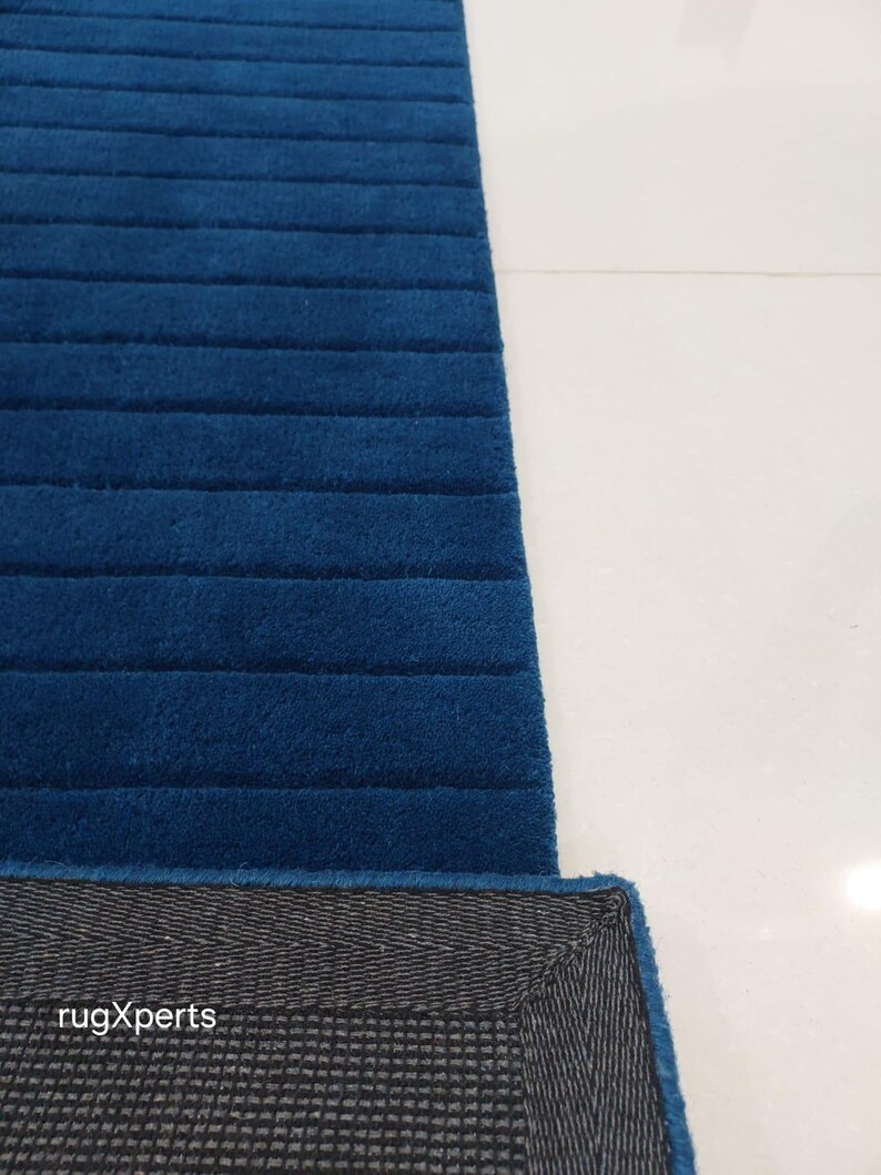 Op de afbeelding: Een gedeelte van een blauw tapijt met een geribbelde textuur. De rand van het tapijt onthult een grijze achterkant met de tekst "rugXperts". Het tapijt ligt op een wit oppervlak.