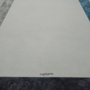 May include: A large rectangular rug with a white central area, framed by sections of grey, brown, and blue. The bottom border is dark grey. The text "rugXperts" is visible near the bottom.
