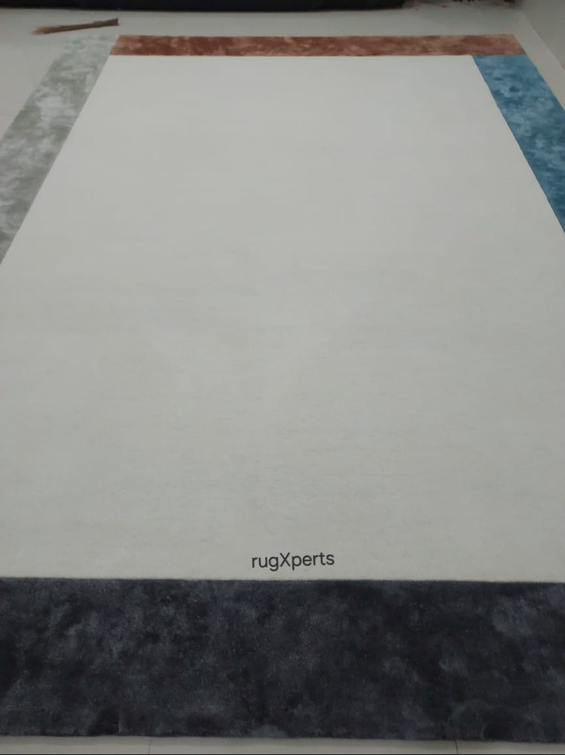 May include: A large rectangular rug with a white central area, framed by sections of grey, brown, and blue. The bottom border is dark grey. The text "rugXperts" is visible near the bottom.