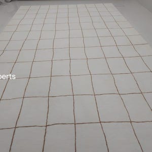 May include: White rug with a grid pattern of brown lines. The rug is made of a soft, plush material and has a subtle sheen.