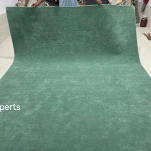 May include: A large, rolled-out, solid-coloured rug in a muted teal green. The rug is made of a soft, plush material and is displayed against a white wall and floor. The text "rugXperts" is visible in the lower left corner.