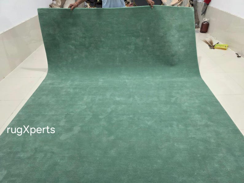 May include: A large, rolled-out, solid-coloured rug in a muted teal green. The rug is made of a soft, plush material and is displayed against a white wall and floor. The text "rugXperts" is visible in the lower left corner.