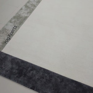 May include: A large, rectangular rug with a cream-coloured central field. The rug features a dark grey border and a lighter grey accent stripe. The words "rugXperts" are printed on the rug. The rug is on a white floor.