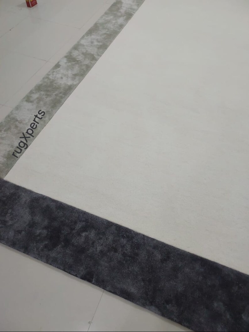 May include: A large, rectangular rug with a cream-coloured central field. The rug features a dark grey border and a lighter grey accent stripe. The words "rugXperts" are printed on the rug. The rug is on a white floor.