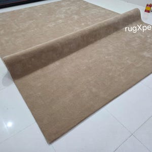 May include: A large, rectangular, beige rug with a rolled edge. The rug is made of a soft, plush material and is displayed on a white tiled floor. The text "rugXperts" is visible on the rug.