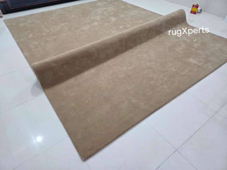 May include: A large, rectangular, beige rug with a rolled edge. The rug is made of a soft, plush material and is displayed on a white tiled floor. The text "rugXperts" is visible on the rug.