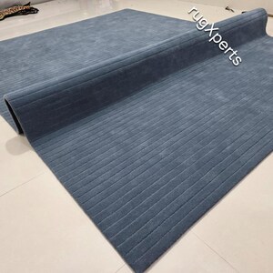 Op de afbeelding: Een grote, opgerolde, blauwe vloerkleed met een geribbelde textuur. Het vloerkleed is gedeeltelijk uitgerold en onthult het zachte oppervlak en de subtiele variaties in de blauwe tint. Het vloerkleed ligt op een lichtgekleurde tegelvloer.