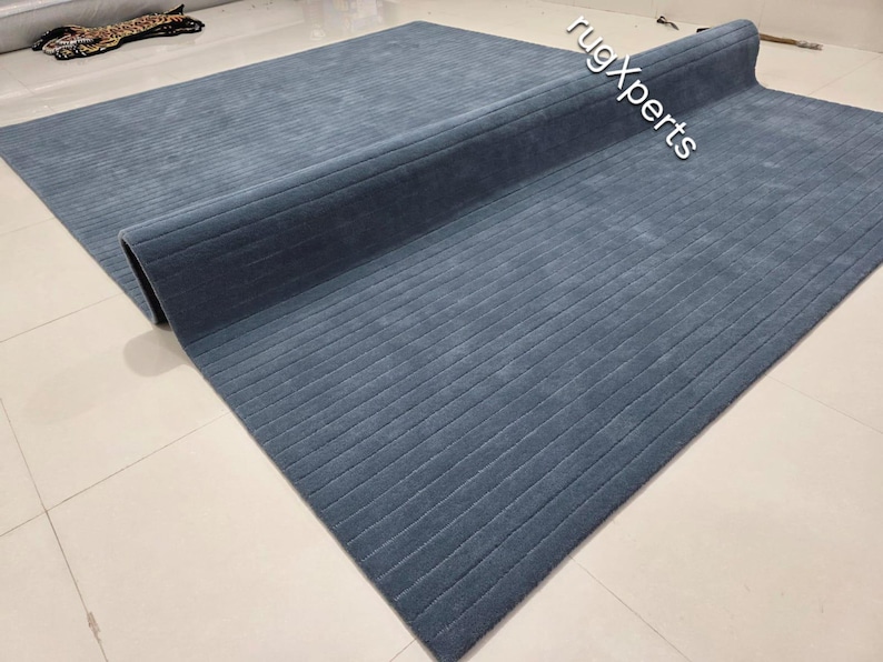 Op de afbeelding: Een grote, opgerolde, blauwe vloerkleed met een geribbelde textuur. Het vloerkleed is gedeeltelijk uitgerold en onthult het zachte oppervlak en de subtiele variaties in de blauwe tint. Het vloerkleed ligt op een lichtgekleurde tegelvloer.