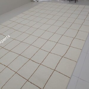 May include: A white rug with a grid pattern of brown lines. The rug is made of wool and has a soft, luxurious feel.