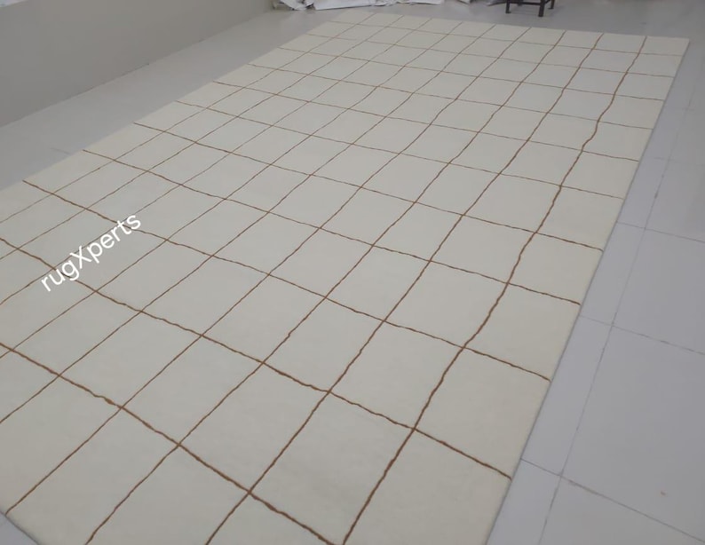 May include: A white rug with a grid pattern of brown lines. The rug is made of wool and has a soft, luxurious feel.