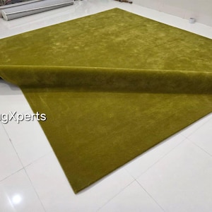 May include: A large, rectangular olive green rug with a rolled edge. The rug is made of a soft, plush material and is displayed on a white tiled floor. The text "rugXperts" is visible in the lower left corner.