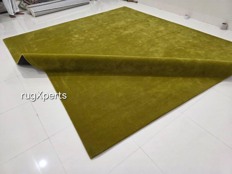 May include: A large, rectangular olive green rug with a rolled edge. The rug is made of a soft, plush material and is displayed on a white tiled floor. The text "rugXperts" is visible in the lower left corner.