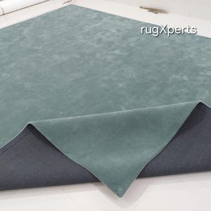 May include: A large, rectangular rug in a muted teal color. The rug's surface has a soft, textured appearance, with a darker gray backing visible where the edge is folded over. The brand name "rugXperts" is visible in the upper right corner.