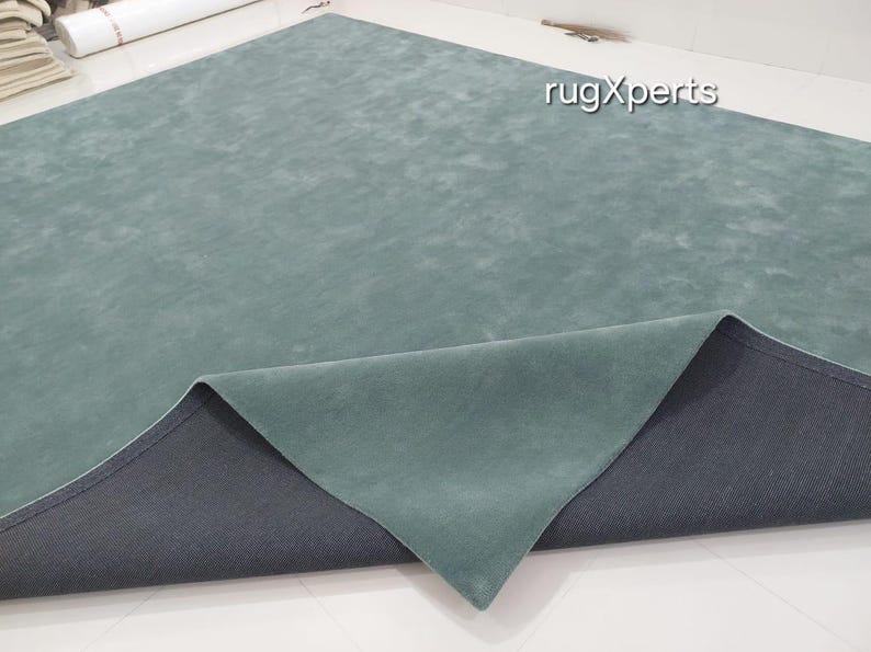 May include: A large, rectangular rug in a muted teal color. The rug's surface has a soft, textured appearance, with a darker gray backing visible where the edge is folded over. The brand name "rugXperts" is visible in the upper right corner.