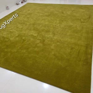 May include: A large, rectangular, olive-green rug. The rug has a plush, textured surface and is placed on a white tiled floor. The words "rugXperts" are visible in the upper left corner.