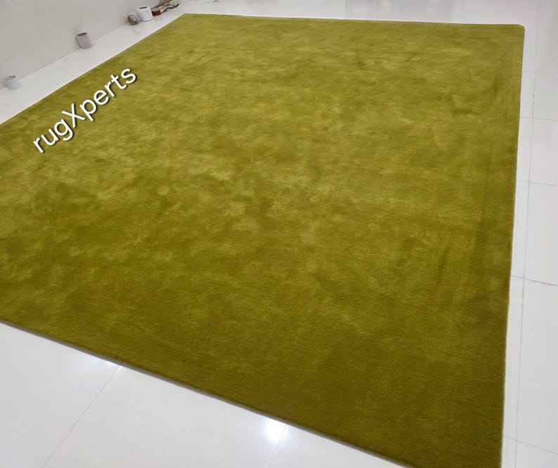 May include: A large, rectangular, olive-green rug. The rug has a plush, textured surface and is placed on a white tiled floor. The words "rugXperts" are visible in the upper left corner.