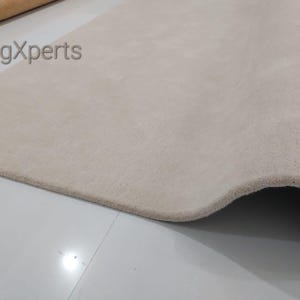 May include: A close-up of a beige rug with a soft, textured surface. The rug is partially rolled, revealing its thickness and construction. The text "rugXperts" is visible in the upper left corner.